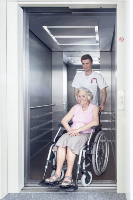 Ein Pfleger schiebt eine ältere Dame im Rollstuhl aus einem Aufzug