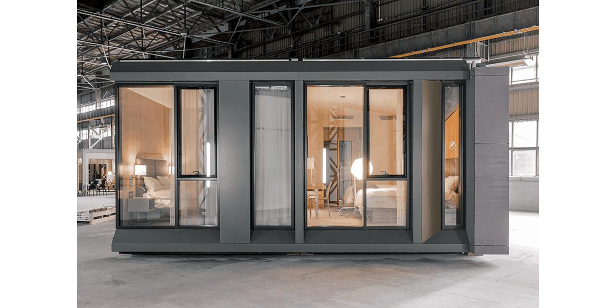 Vorgefertigte Kapsel-Suite. Komplett möbliert, mit warmen Elementen. Die Außenwand ist grau und hat viele Fenster.