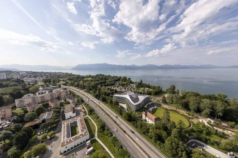 Vogelaufnahme des Gebäude des IOC. Im Hintergrund befindet sich der Genfer See, der Himmelst leicht wolkig, aber sonnig.____