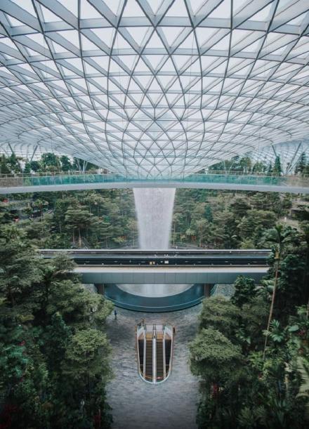 Indoor Wasserfall, der aus einem Glasdach entspringt. Um den Wasserfall herum gibt es grüne Pflanzen und zwei Brücken.