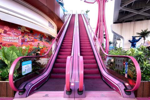 Breite Ansicht einer pinken Rolltreppe in einem Einkaufszentrum mit grünen Pflanzen.